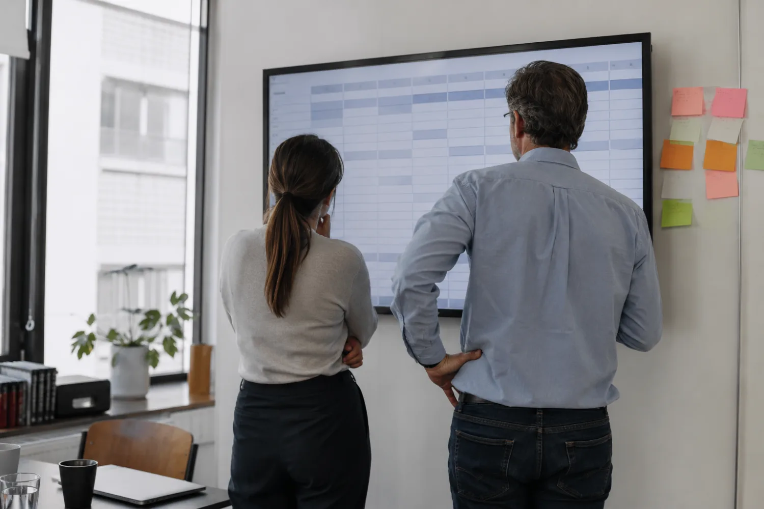 Dos personas de espaldas revisan una tabla de datos en una pantalla grande, con notas de colores pegadas al lado