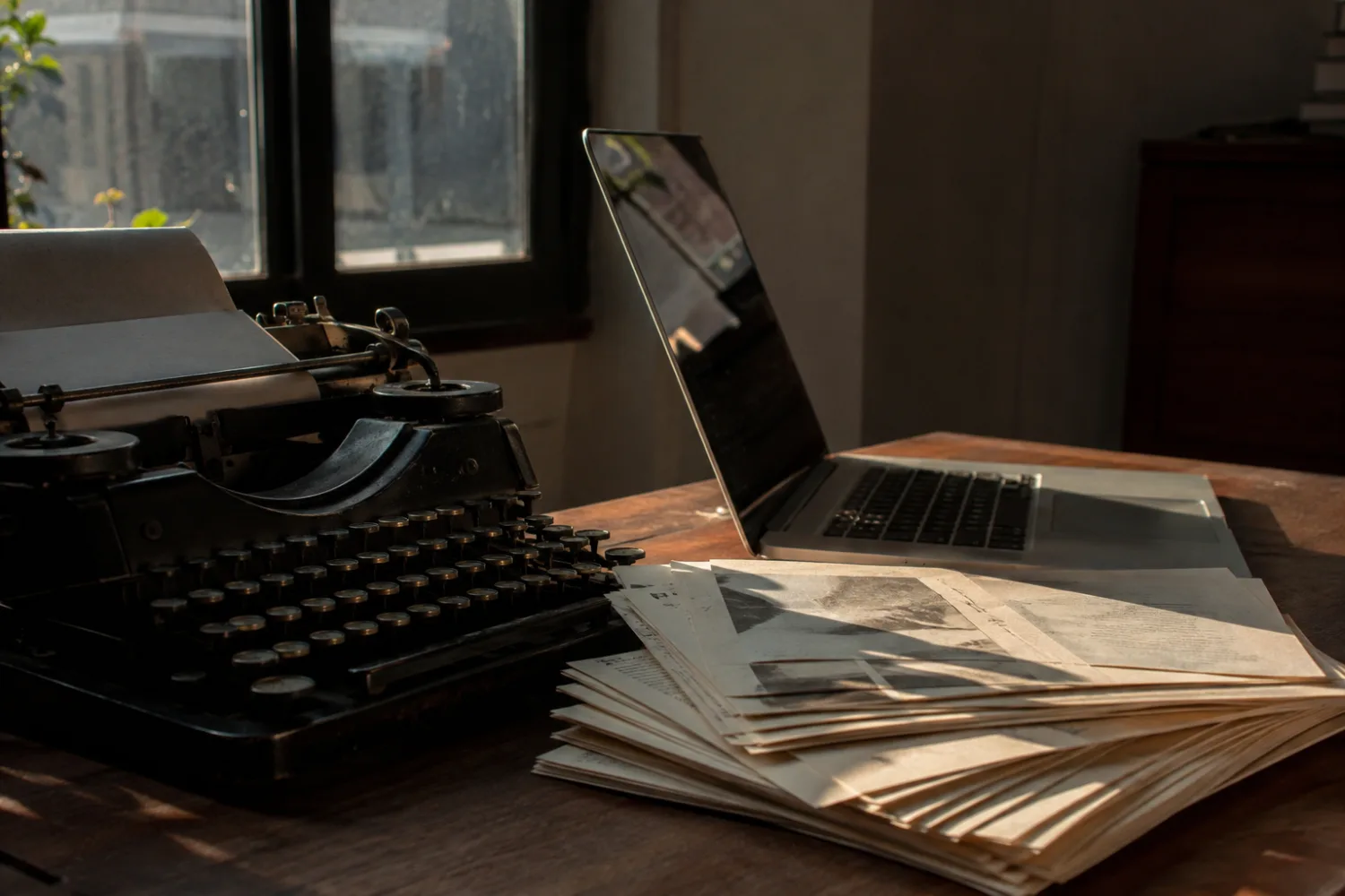 Una máquina de escribir antigua junto a un portátil abierto, con una pila de folios entre las dos